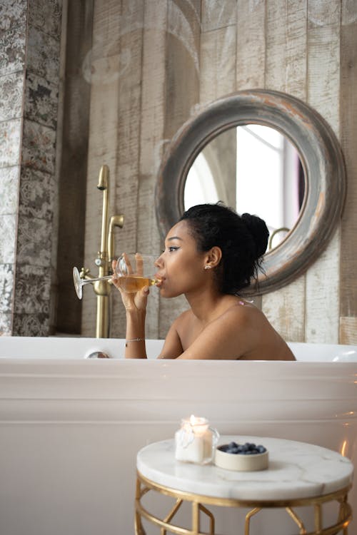 Side view of young ethnic female sitting in bathtub and drinking wine while enjoying spa procedure in bathroom