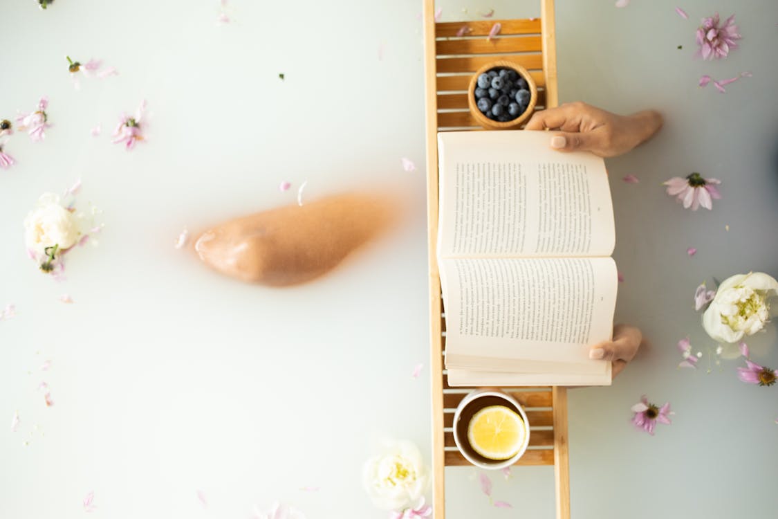 Top view of crop unrecognizable lady in white water in bathtub with fresh colorful flower petals with wooden tray with cup of tea with lemon and blueberries while reading book