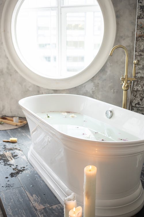 Bathtub with white water and fresh colorful flower petals with tap next to burning wax candles on floor and window on wall in light apartment