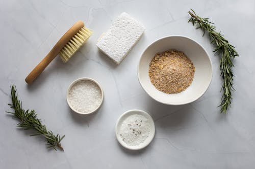 Top view of bowls with ingredients for organic scrub arranged with pumice stone and fresh branches of rosemary