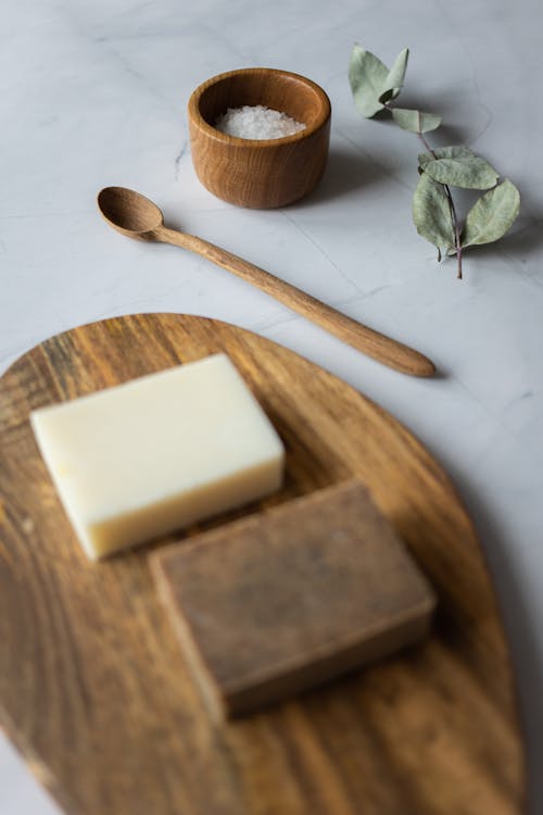 From above of aromatic rectangular shaped handmade soap placed on wooden board near scrub with salt and rosemary