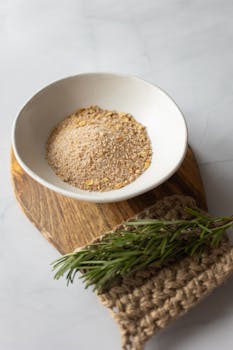 From above of white bowl filled with powder of oat flakes near fresh verdant branch of rosemary on timber board