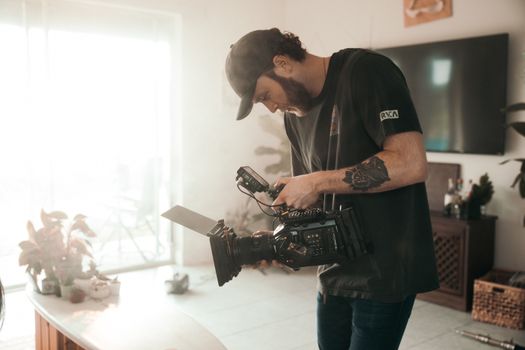 Caucasian male filmmaker setting up camera indoors with bright natural light.