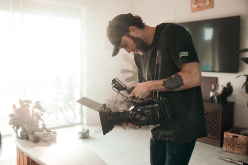 Free Caucasian male filmmaker setting up camera indoors with bright natural light. Stock Photo