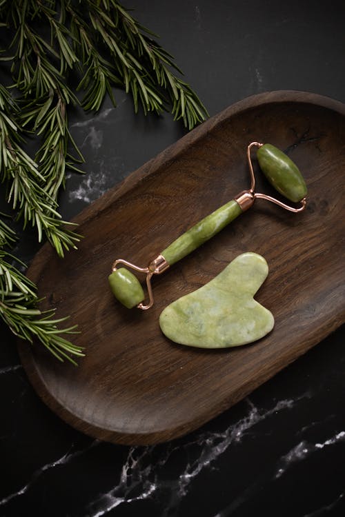 Free From above of Jade roller and Gua Sha massage tool placed on wooden plate on black marble table with fresh twigs of rosemary Stock Photo