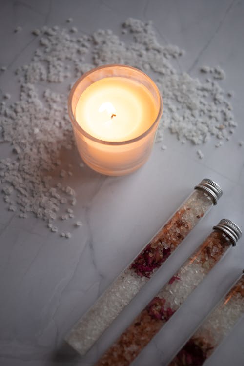 High angle of jars with colorful natural salt for bath placed near burning candle on white marble table