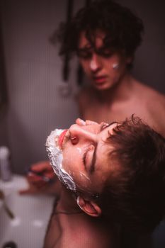 Two shirtless men share a tender shaving moment in a cozy bathroom setting.