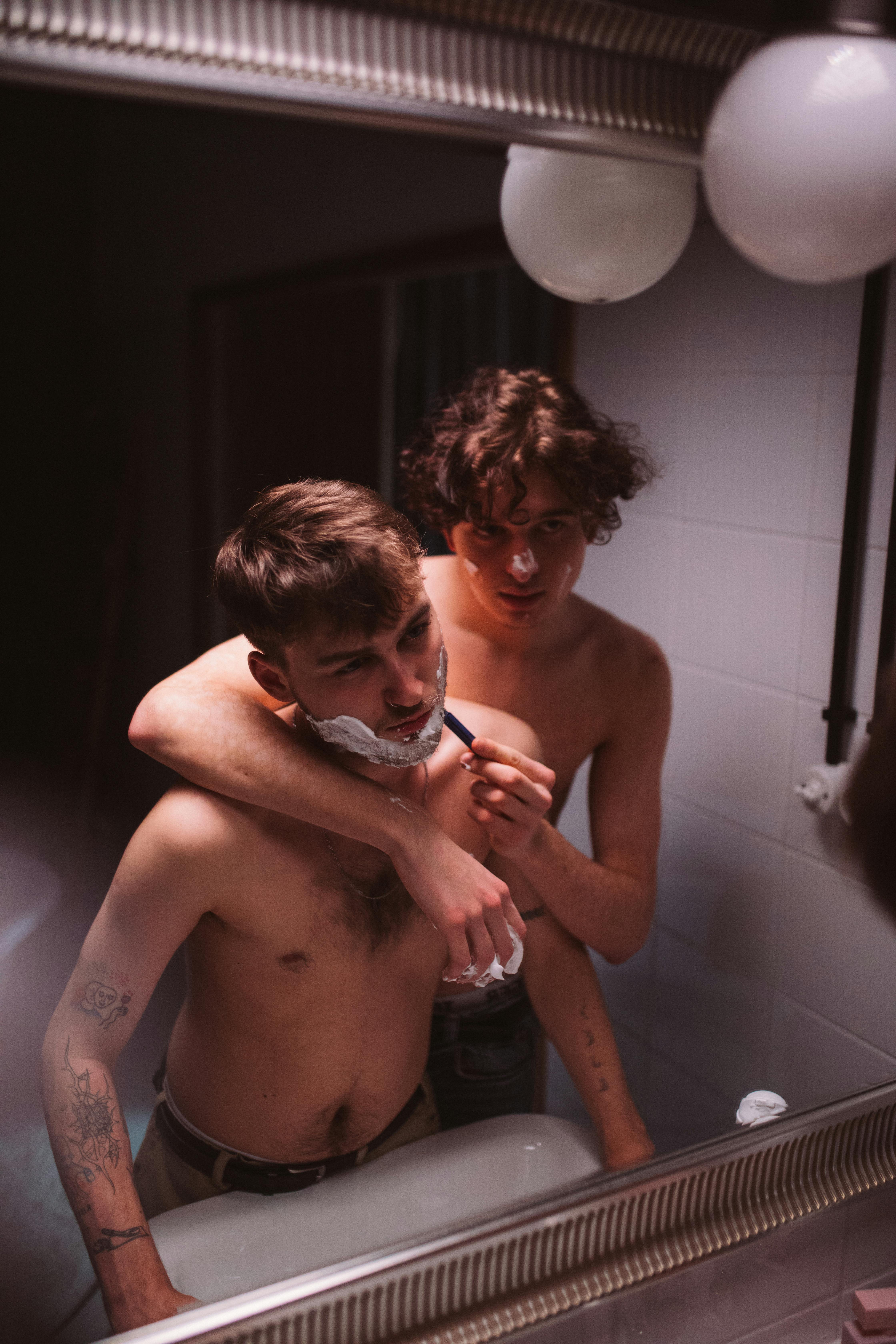 A candid moment of affection between two shirtless men in a bathroom, one assisting the other with shaving.