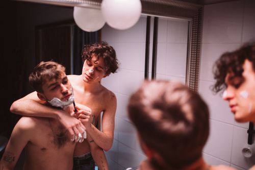 Free A romantic moment captured where two shirtless young men are shaving together in front of a mirror showcasing love and trust. Stock Photo