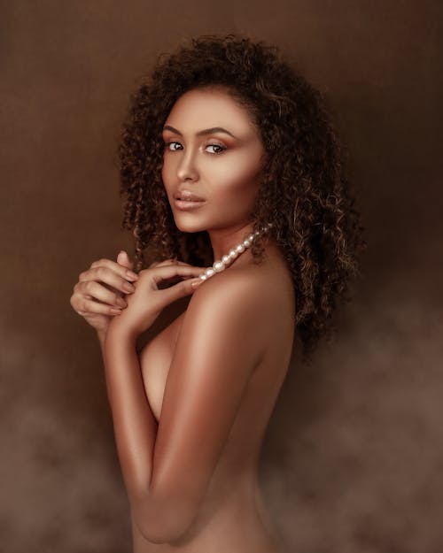 Side view of nude African American female touching necklace standing in smoke in studio and looking at camera