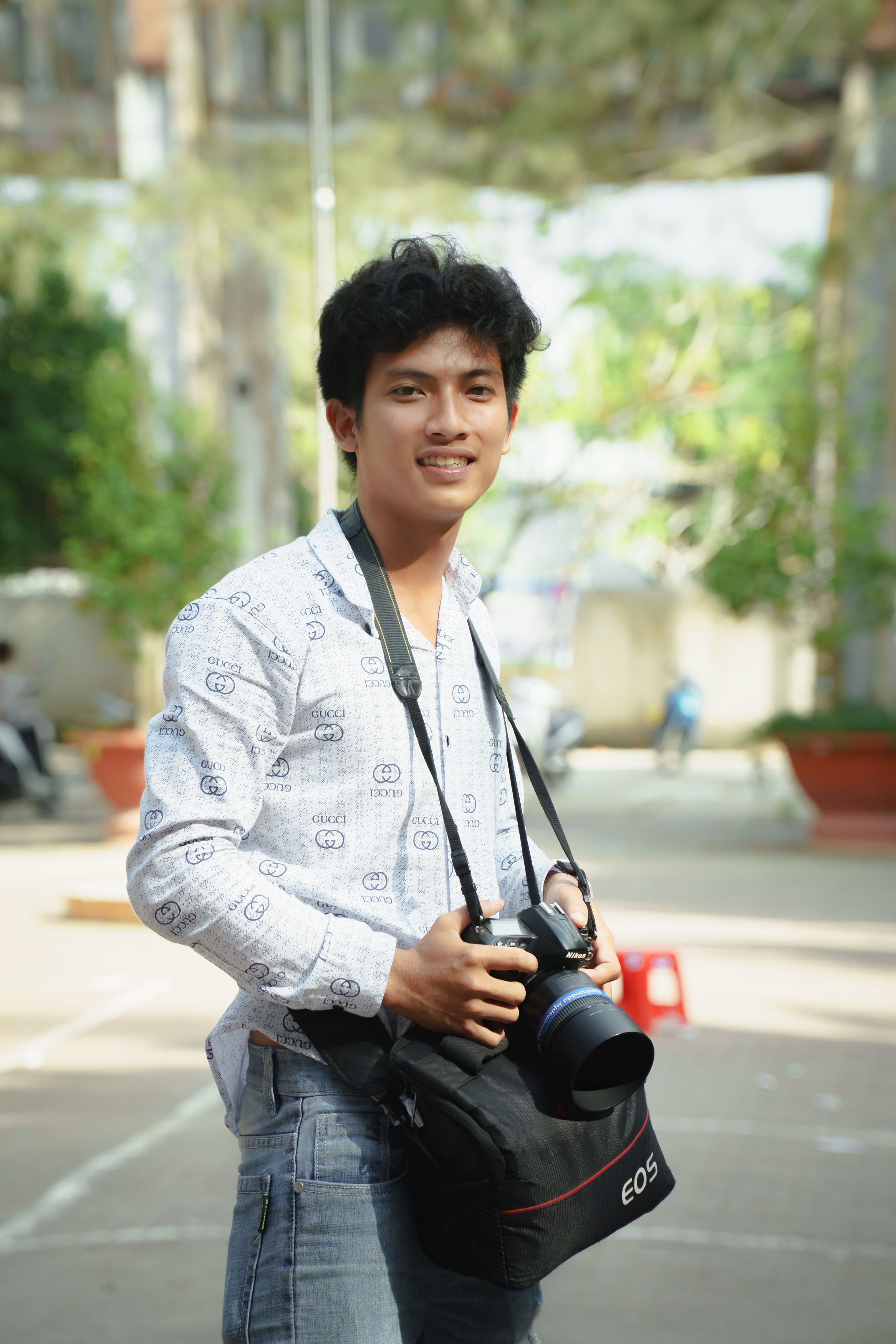Young man holding a camera, smiling outdoors in daylight.