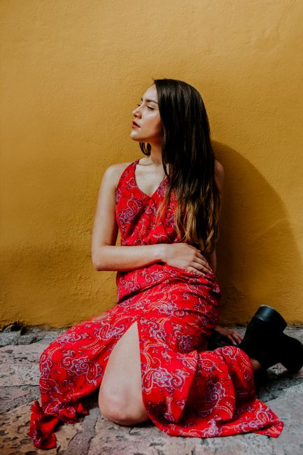 Stylish woman in a red dress sitting against a vibrant yellow wall, showcasing a modern fashion look.