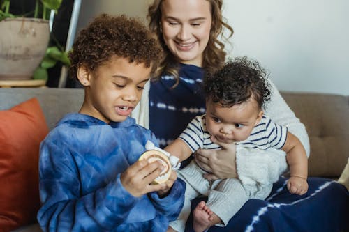 Free Positive mum hugging curios African American baby reaching hands to sibling playing with toy at home Stock Photo