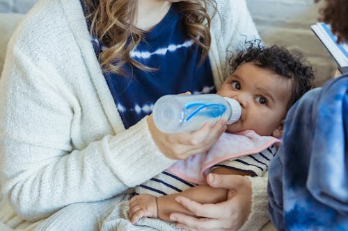 Free High angle of crop mother embracing little African American baby and feeding from bottle sitting on sofa near son Stock Photo