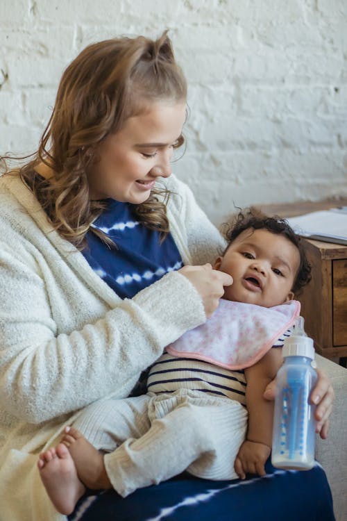 Free Cheerful mother looking at cute barefoot African American child while sitting on couch with feeding bottle in room at home Stock Photo
