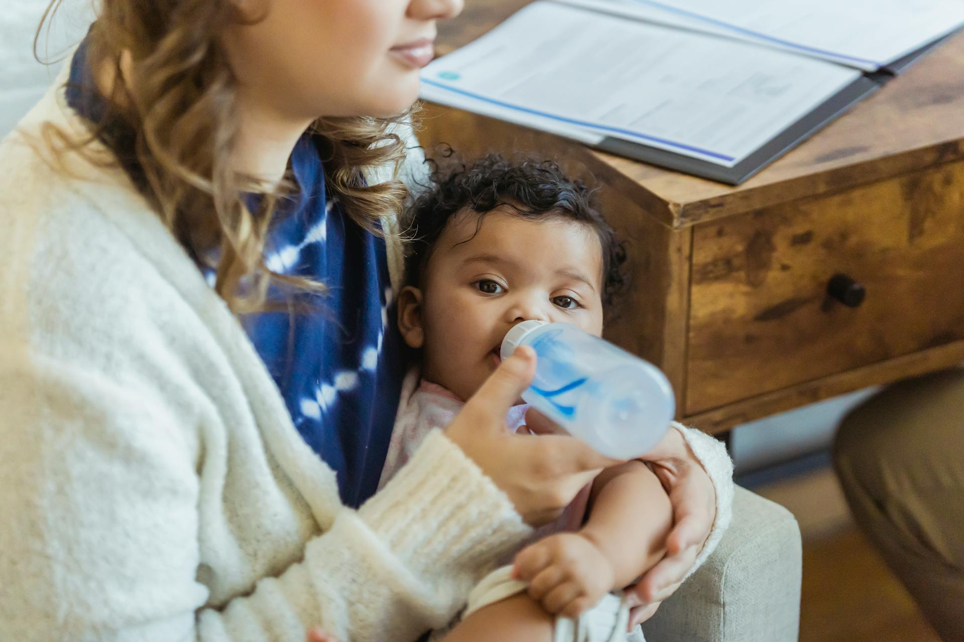 Unrecognizable mother bottle feeding cute little African American baby in hands while sitting near wooden drawer in light room at home