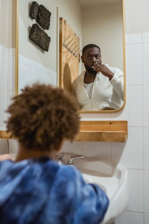 Focused African American male brushing teeth and looking at reflection of mirror while standing in bathroom with unrecognizable son during daily routine