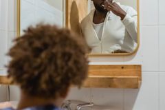 Focused African American male brushing teeth and looking at reflection of mirror while standing in bathroom with unrecognizable son during daily routine