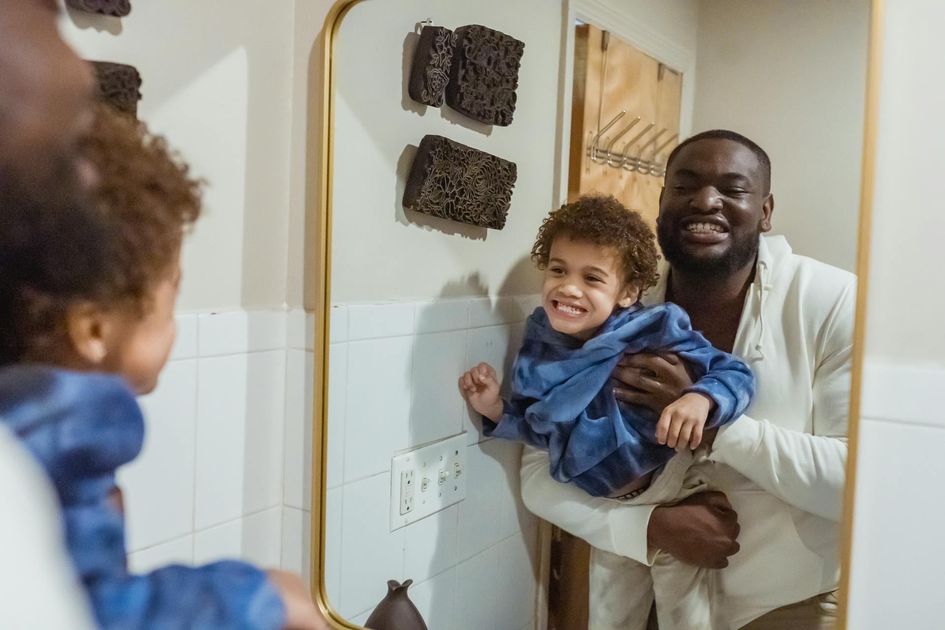 Content African American father with son in hands looking at reflection of mirror and showing teeth while standing in bathroom