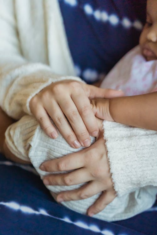 Unrecognizable gentle mother in warm wear hugging crop cute little African American baby in hands while sitting in room at home
