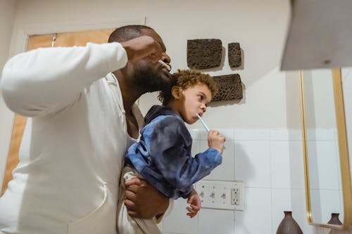 Side view of focused African American father with boy in hands brushing teeth with toothbrushes during morning routine in bathroom