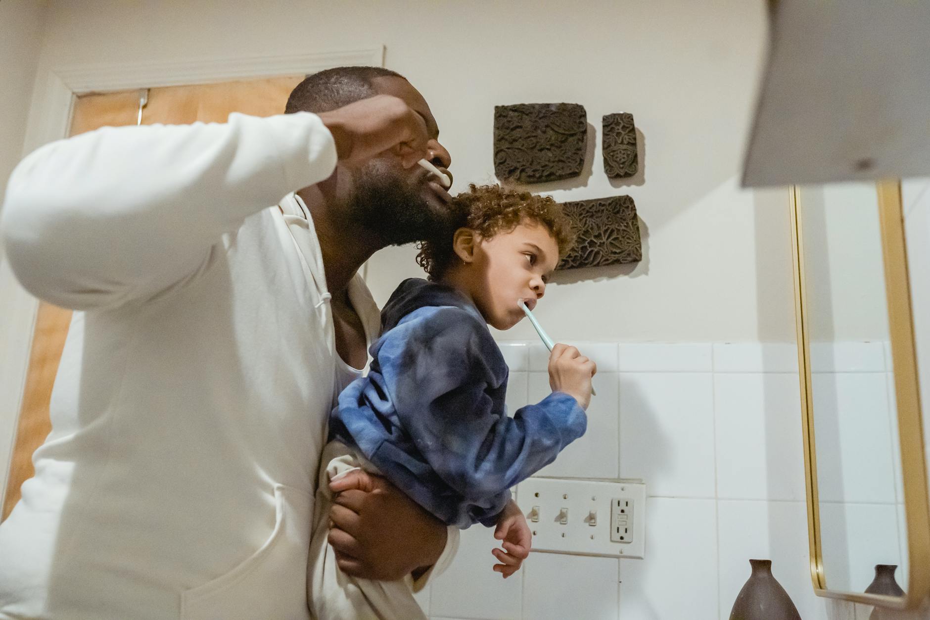 Side view of focused African American father with boy in hands brushing teeth with toothbrushes during morning routine in the bathroom.