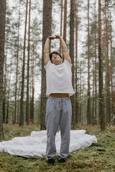 A man stretches in serene forest scenery, standing on a bed setup outdoors.