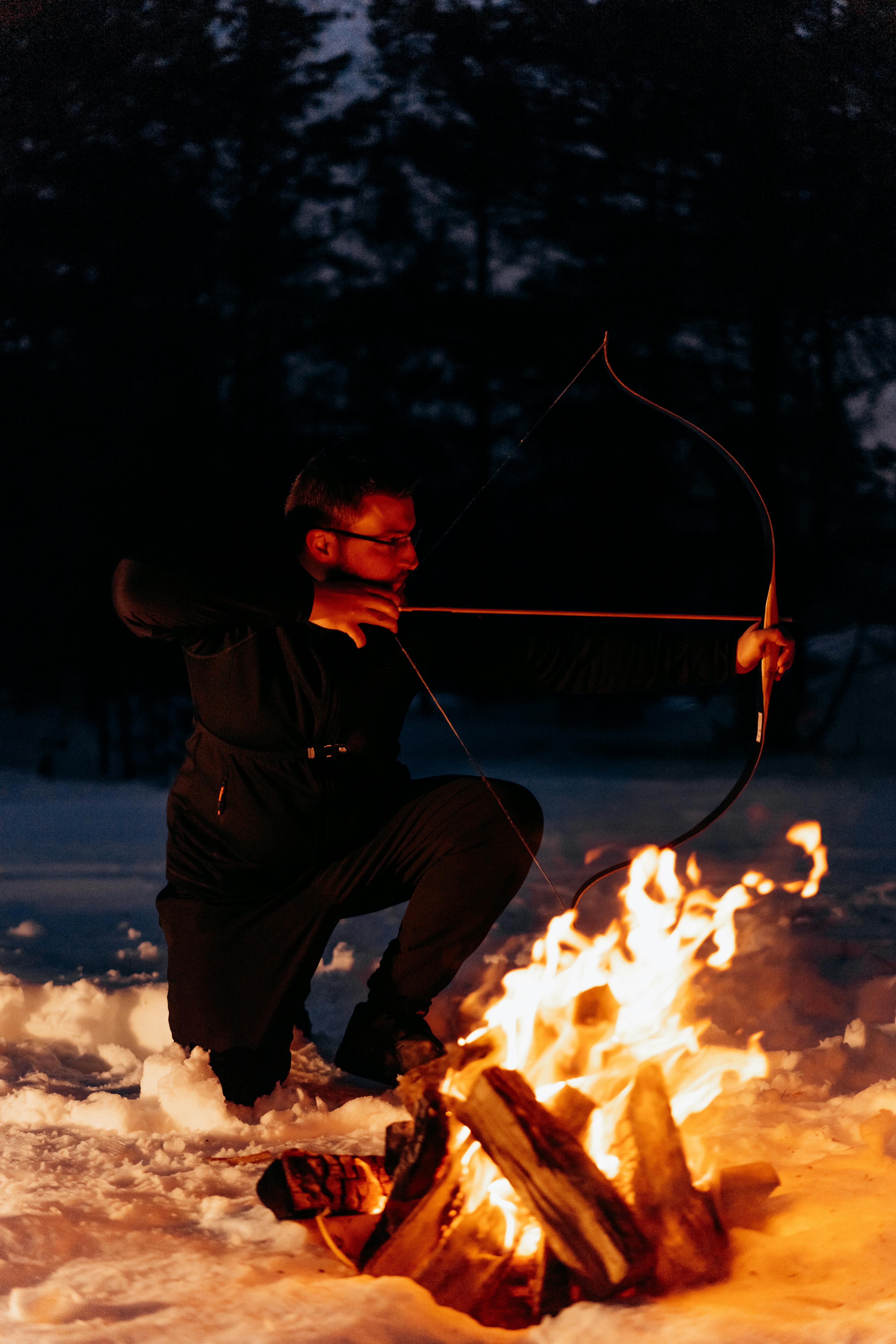 A man with a bow and arrow kneels by a campfire in a snowy forest during winter night.