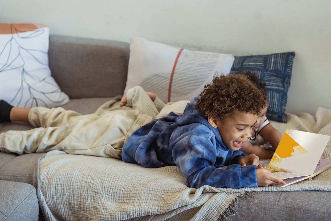 Side view of smiling African American boy with anonymous baby reading book while lying on comfortable sofa in cozy room