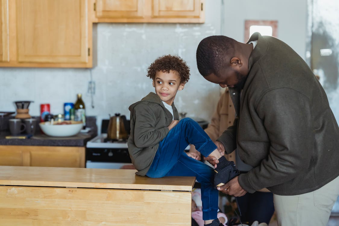 Focused African American father helping cheerful black son to put on footwear while sitting on wooden table in modern kitchen