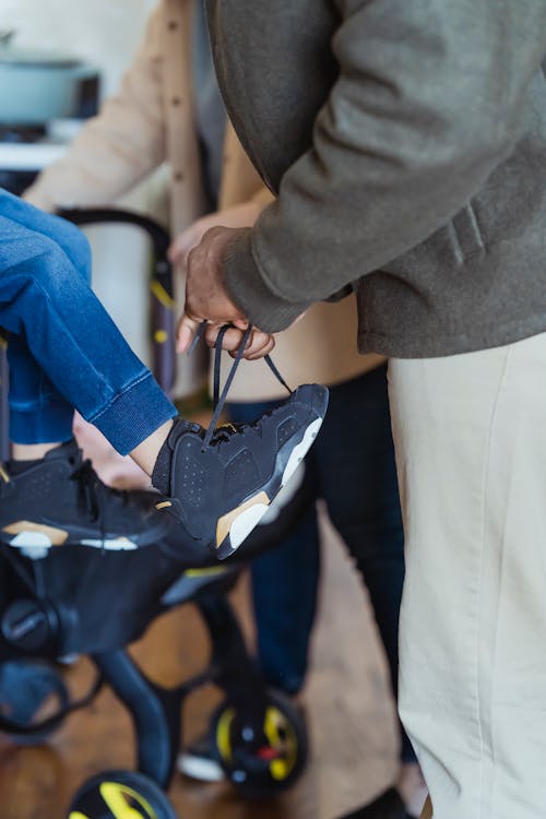 Unrecognizable father helping son in jeans to put on footwear while sitting in light apartment with baby stroller at home