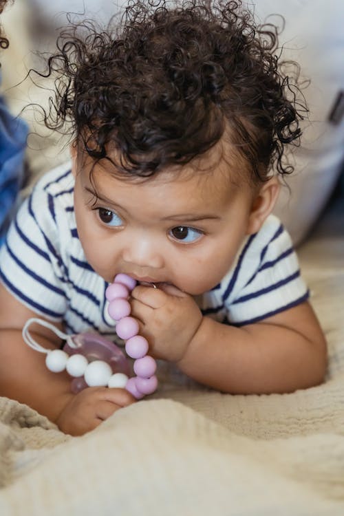 Cute little African American baby with curly hair lying on sofa while playing with beads in room on blurred background