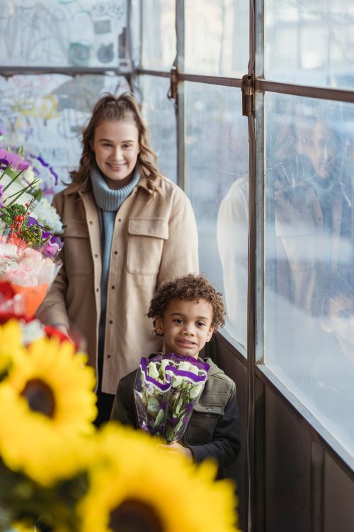 Free Cheerful mother and African American boy with bouquet of flowers looking away while standing near windows in modern floral store Stock Photo