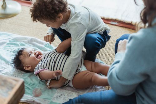Free Happy ethnic boy in casual clothes playing with laughing cute infant lying on floor at home Stock Photo