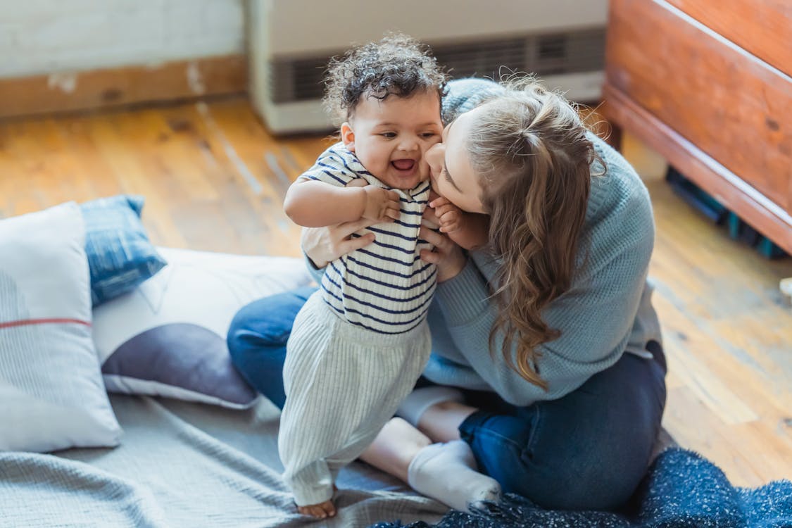 Free Full length caring mother in casual wear hugging and kissing cute ethnic baby cheek while sitting on floor in light living room Stock Photo