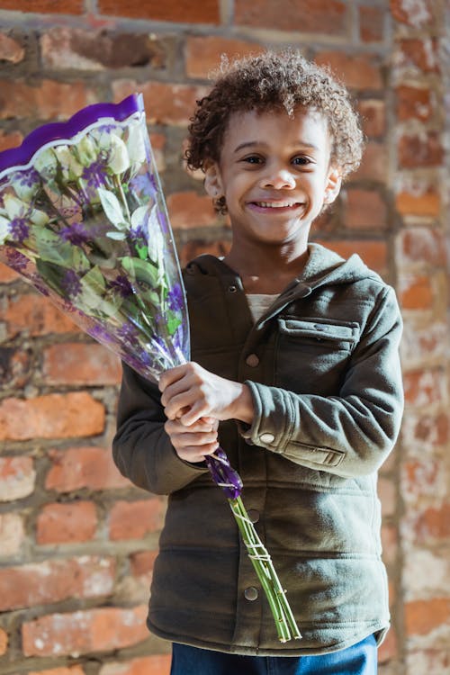 Happy cute African American boy in casual clothes holding bouquet of fragrant white flowers and looking at camera with smile while standing against shabby brick wall