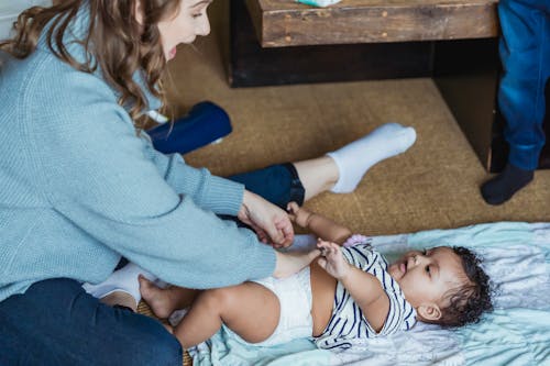 Free From above excited young mother changing diaper and playing with adorable ethnic baby lying on linen on floor Stock Photo