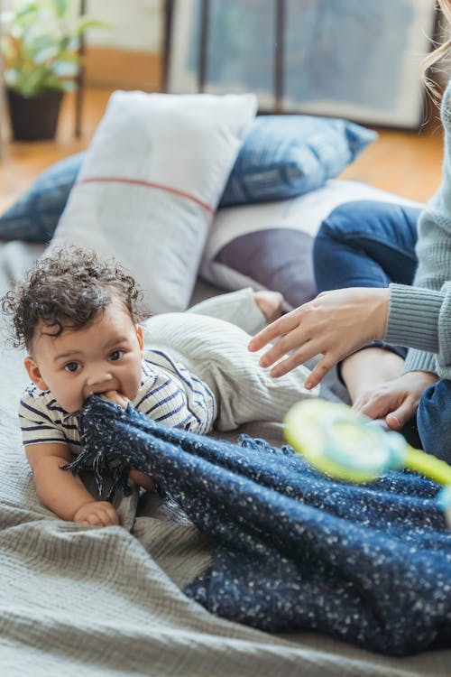 Free Adorable ethnic baby biting plaid and looking at camera while lying on belly on floor at home Stock Photo