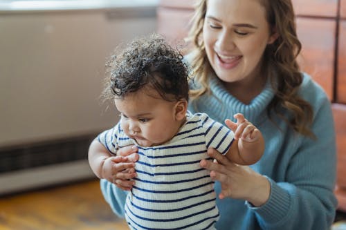 Crop cheerful mom with mouth opened teaching curious African American baby walking at home