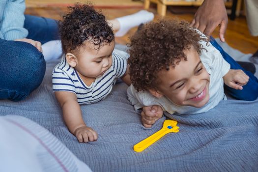 Crop African American dad tickling laughing son lying on floor near curious baby near mother