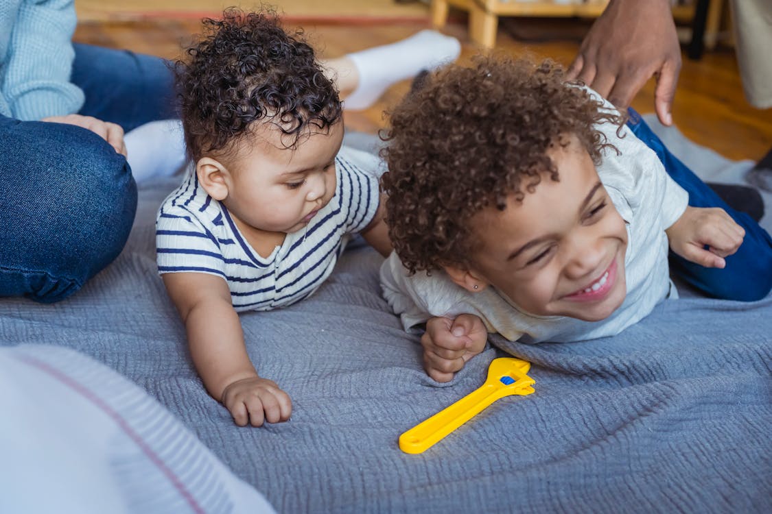 Crop African American dad tickling laughing son lying on floor near curious baby near mother