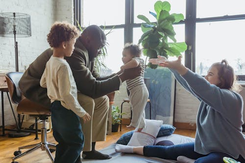 Free Side view of joyful mother with raised hands on floor opposite African American dad lifting baby while sitting near son Stock Photo