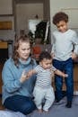 Happy mom sitting on floor and teaching baby to stand on feet and elder kid looking at sibling