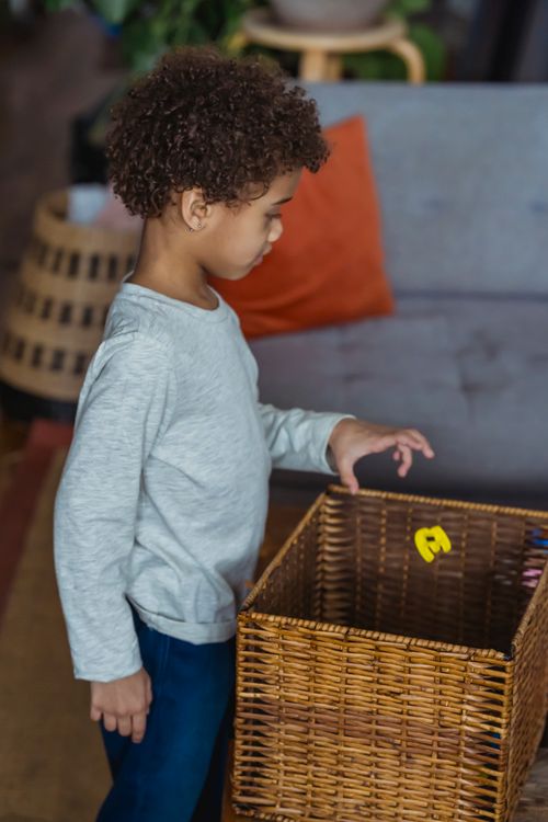 Free Side view of little child in jeans standing in cozy room with hand raised over wicker basket and looking down Stock Photo