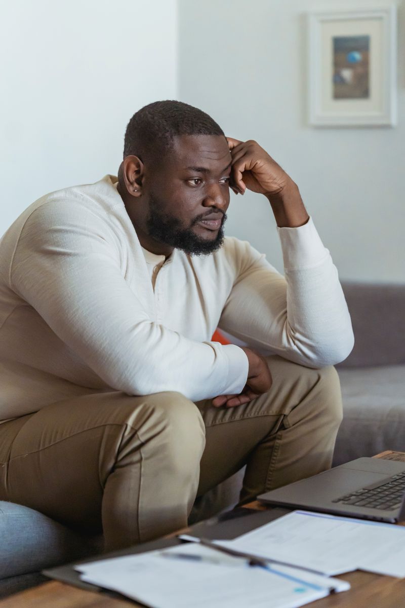 A Black man sits at a low table with a laptop and paperwork, hand resting against his head, looking drained and unfocused while trying to work.