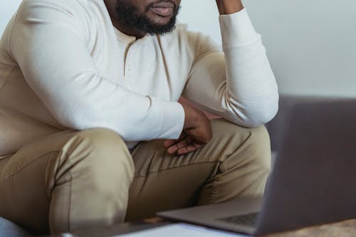 Crop anonymous black male freelancer in casual clothes looking away while sitting and working near laptop