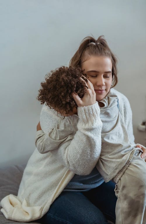 Tender woman with closed eyes embracing loving kid while sitting on sofa and calming down