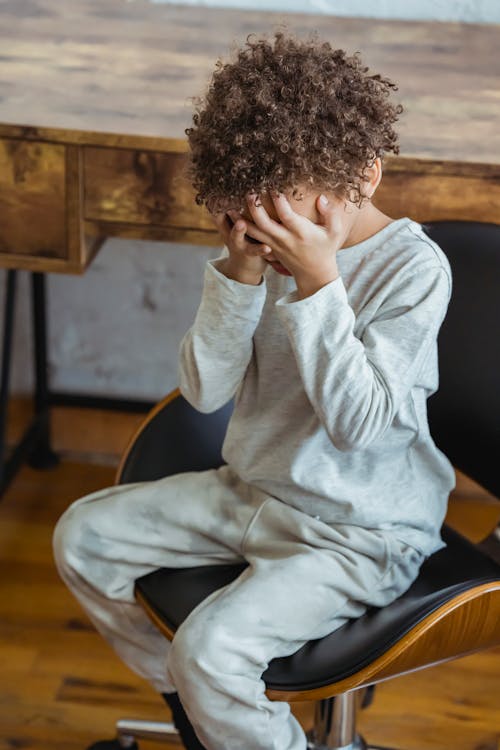 Free From above of crying African American child with curly hair wearing casual clothes sitting in chair near table and covering eyes with hands Stock Photo
