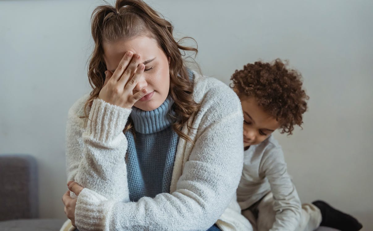 Free Frustrated mother with hand on forehead and closed eyes sitting near African American son near wall in room at home Stock Photo