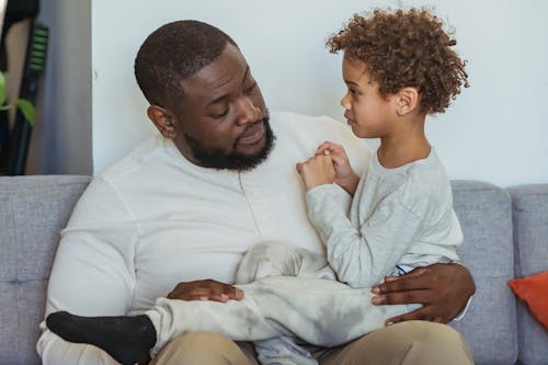 Content African American boy sitting on knees of caring black bearded father on couch in light living room at home